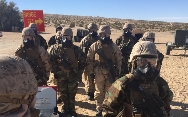 A group of military personnel in full CBRN (Chemical, Biological, Radiological, Nuclear) protective gear including gas masks and MOPP suits are assembled in a desert training environment. A sign reading 'RANGE 225' with a directional arrow is visible in the background. The setting appears to be a military training range, likely at a U.S. Marine Corps installation based on the MARPAT camouflage patterns visible.