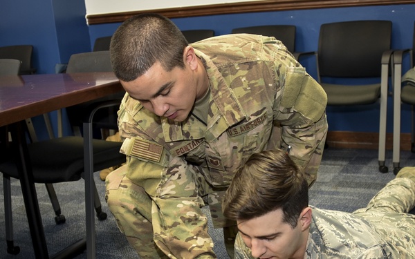 Two U.S. Air Force personnel are conducting CPR/first aid training in what appears to be a classroom or briefing room. One service member in OCP (Operational Camouflage Pattern) uniform is supervising while another in ABU (Airman Battle Uniform) practices airway management techniques on a CPR training mannequin. The setting suggests a formal military medical readiness training exercise.