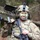 A soldier in full combat gear poses in a wooded environment, wearing MultiCam camouflage uniform and helmet with night vision mount. The soldier has green and black face paint applied and is carrying a shoulder-mounted weapon system labeled 'DUMMY' along with a rifle. The setting appears to be a military training exercise based on the training equipment markings.