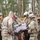 A group of approximately seven to eight soldiers in U.S. Army MultiCam/OCP camouflage uniforms are gathered in a forested area conducting what appears to be a tactical briefing or operations planning session. One soldier, wearing a patrol cap rather than a helmet, is pointing at a document or map being held by another soldier. The setting appears to be a deciduous/mixed forest with fallen autumn leaves on the ground, consistent with a field training exercise environment.