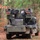 A military vehicle, appearing to be a HUMVEE (HMMWV), is equipped with what appears to be a Avenger air defense missile system mounted on its rear. The vehicle is positioned in a wooded area with autumn foliage on the ground. The system features dual launcher pods with visible sensor/optic housings and a central cooling unit.