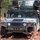 A military HMMWV (Humvee) is positioned in a wooded area covered with autumn fallen leaves. The vehicle is equipped with what appears to be a Avenger air defense missile system mounted on the roof, featuring dual missile pod launchers. Unit markings '1-174 AD' and 'C12' are visible on the front bumper, indicating an Air Defense Artillery unit.