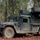 A military HMMWV (Humvee) equipped with what appears to be a missile launcher system, likely an Avenger air defense system or similar SHORAD platform, positioned in a wooded forest environment. The vehicle is covered with camouflage netting over the weapon system mounted on the rear. The setting suggests a field exercise or tactical deployment position among deciduous and coniferous trees.