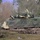 A tracked armored fighting vehicle, consistent with a Bradley Infantry Fighting Vehicle (IFV), is positioned in a muddy woodland clearing. The vehicle is painted in desert tan/sand coloring and is draped with green camouflage netting over its hull and turret. The setting appears to be a military exercise or field operation in a temperate European-style forest environment.