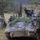 Military personnel are operating armored tracked vehicles, likely M113 Armored Personnel Carriers (APCs), in a wooded training area. The vehicles are adorned with natural foliage camouflage and carry crew members in the open hatches. At least two soldiers on foot are visible near the treeline observing or directing the exercise.