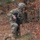 A soldier in full combat gear is moving through a wooded area covered in autumn foliage. The soldier is in a crouched, tactical movement posture while carrying a rifle, suggesting a field training exercise or patrol operation. The environment is characterized by fallen orange-brown leaves and bare deciduous trees.