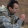 A male soldier wearing US Army ACU (Army Combat Uniform) digital camouflage pattern is seated outdoors, appearing to eat or drink something from a small package or pouch. He is equipped with a tactical rucksack and load-bearing gear. The setting appears to be a field training environment with sparse grass and dirt ground.