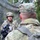 Two military personnel in MultiCam/OCP camouflage uniforms are engaged in what appears to be a media interview or documentation session in a field environment. A soldier facing the camera is being filmed or interviewed, while another soldier with the name tape 'SMITH' visible on their cap stands with their back to the viewer holding a video camera. The setting includes camouflage netting strung between trees, suggesting a tactical field environment.