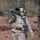A soldier in full combat gear walks through a wooded area with autumn leaf coverage on the ground. The soldier is carrying a man-portable air-defense system (MANPADS) on their shoulder, likely an FIM-92 Stinger or similar missile launcher, along with an assault rifle. The soldier's face is painted with green camouflage paint, suggesting a field training exercise or operational deployment.