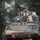 A military armored fighting vehicle, consistent with an M2 Bradley Infantry Fighting Vehicle, is photographed in a forested environment. The vehicle is heavily covered with camouflage netting draped over its turret and hull, and shows signs of mud and field use. The image is taken from a front-facing angle, capturing the vehicle's turret, main gun, and forward hull section among pine trees.