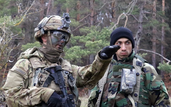 Two military personnel are standing in a wooded outdoor area, appearing to be engaged in a briefing or coordination activity. The soldier on the left is in full combat gear with camouflage face paint and is pointing in a direction, while the soldier on the right wears a different camouflage pattern and a black beanie. The setting and differing uniforms suggest a joint training exercise or multinational military operation.
