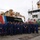 A group of approximately 30 United States Coast Guard personnel pose for a formal group photograph on a dock beside a Coast Guard vessel. The crew members are dressed in matching navy blue uniforms and caps. The ship displays the distinctive USCG diagonal stripe livery in red, white, and blue, along with the official Coast Guard seal dated 1790.
