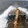 An aerial or elevated view of a research or offshore support vessel's working deck taken during rough sea conditions. Large waves are washing over the bow and port side of the vessel, indicating significant swell. The deck is equipped with heavy industrial crane/winch machinery painted in yellow/orange, consistent with oceanographic or subsea operations.
