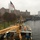 The image shows the bow of the USCGC Abbie Burgess, a United States Coast Guard buoy tender designated as a 'Downeast Tender,' docked at a waterfront facility on an overcast, rainy day. An American flag flies at what appears to be half-staff from a tall flagpole on shore, with urban residential buildings visible in the background. A gray naval vessel bearing the number '4' is docked nearby to the right.