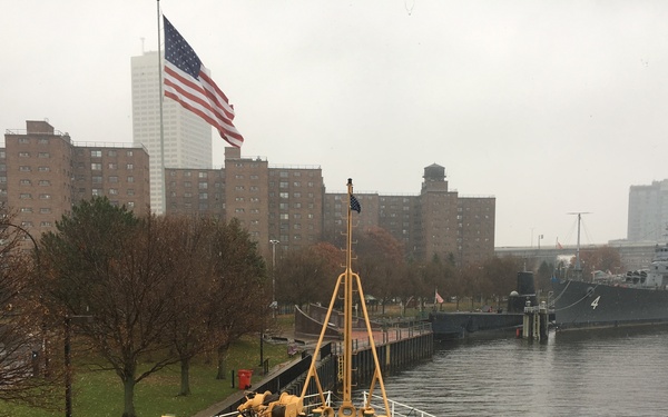 The image shows the bow of the USCGC Abbie Burgess, a United States Coast Guard buoy tender designated as a 'Downeast Tender,' docked at a waterfront facility on an overcast, rainy day. An American flag flies at what appears to be half-staff from a tall flagpole on shore, with urban residential buildings visible in the background. A gray naval vessel bearing the number '4' is docked nearby to the right.