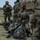 A group of military personnel in camouflage uniforms are lined up on a grassy field with their equipment and rifles stacked on the ground beside them. One soldier in the foreground appears to be reviewing a document or map while others stand in formation. The scene suggests a pre-mission briefing, equipment inspection, or staging area during a training exercise or operational deployment.
