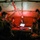 A group of military personnel are gathered inside what appears to be a tactical field tent or command post during nighttime operations. They are clustered around a table with a laptop computer, illuminated by a single red light hanging from the tent structure. The scene suggests a briefing, planning session, or operational coordination meeting.