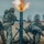 The image shows a group of soldiers in camouflage uniforms operating a mortar system that is actively firing, with a visible muzzle blast and flame at the top of the tube. The soldiers are positioned around the mortar in a field setting under an overcast sky. The scene appears to depict a military training exercise or live-fire operation.
