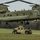 A United States Army CH-47 Chinook helicopter hovers low over a grassy field while soldiers work to attach a tan/beige HMMWV (Humvee) for a sling-load operation. At least two soldiers in camouflage uniforms are visible on and around the Humvee, connecting rigging to the helicopter's underside. The scene appears to be a training exercise conducted in a cleared field surrounded by bare trees.