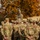 A military leader, seated elevated on what appears to be an armored vehicle, addresses a large formation of soldiers in MultiCam/OCP camouflage uniforms. The soldiers are gathered in a semicircle facing the speaker, suggesting a briefing or after-action review. The setting appears to be an outdoor training area during autumn, indicated by the orange and yellow foliage in the background.