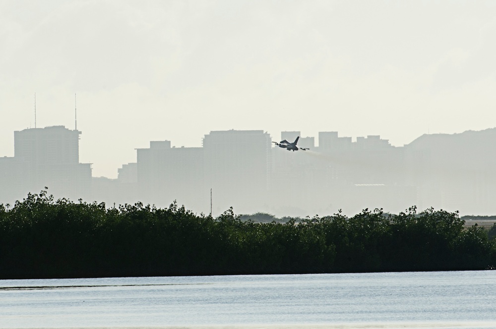 A military jet aircraft is captured in low-level flight against a hazy urban skyline backdrop. The scene is photographed from across a body of water, with a dense treeline of what appears to be mangroves in the middle ground. The overall image has a muted, atmospheric quality due to haze or humidity.