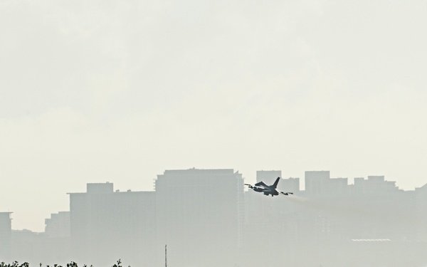 A military jet aircraft is captured in low-level flight against a hazy urban skyline backdrop. The scene is photographed from across a body of water, with a dense treeline of what appears to be mangroves in the middle ground. The overall image has a muted, atmospheric quality due to haze or humidity.