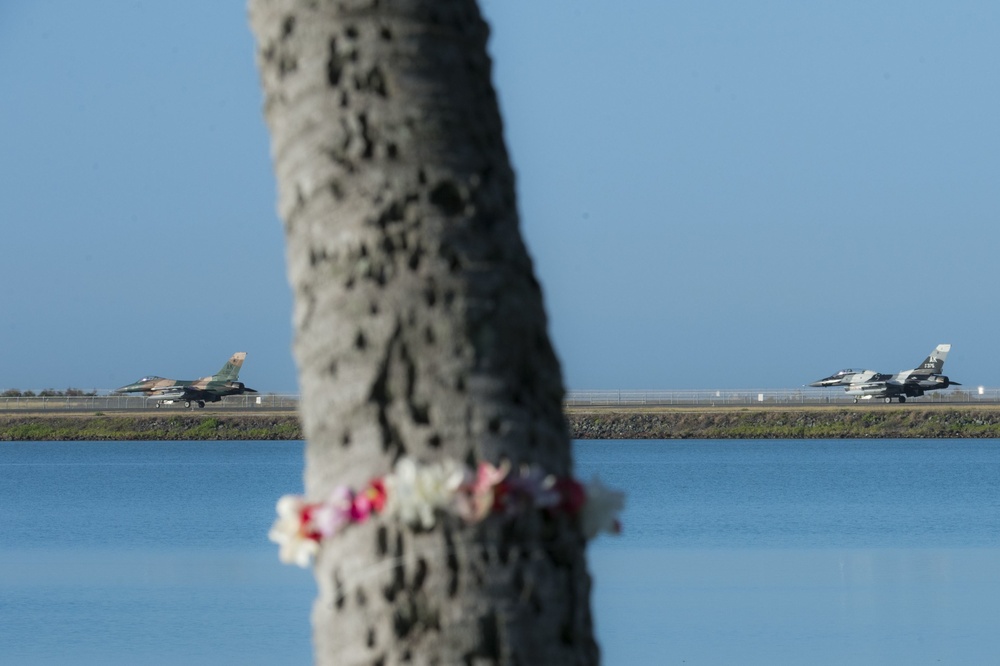 A close-up of a palm tree trunk with flower leis attached to it stands in the foreground, with a calm body of water in the middle ground. In the background, two military fighter jets — one in camouflage green/brown and one in gray — are visible on what appears to be an airfield runway. The composition creates a contrast between the floral memorial element and the military aircraft.