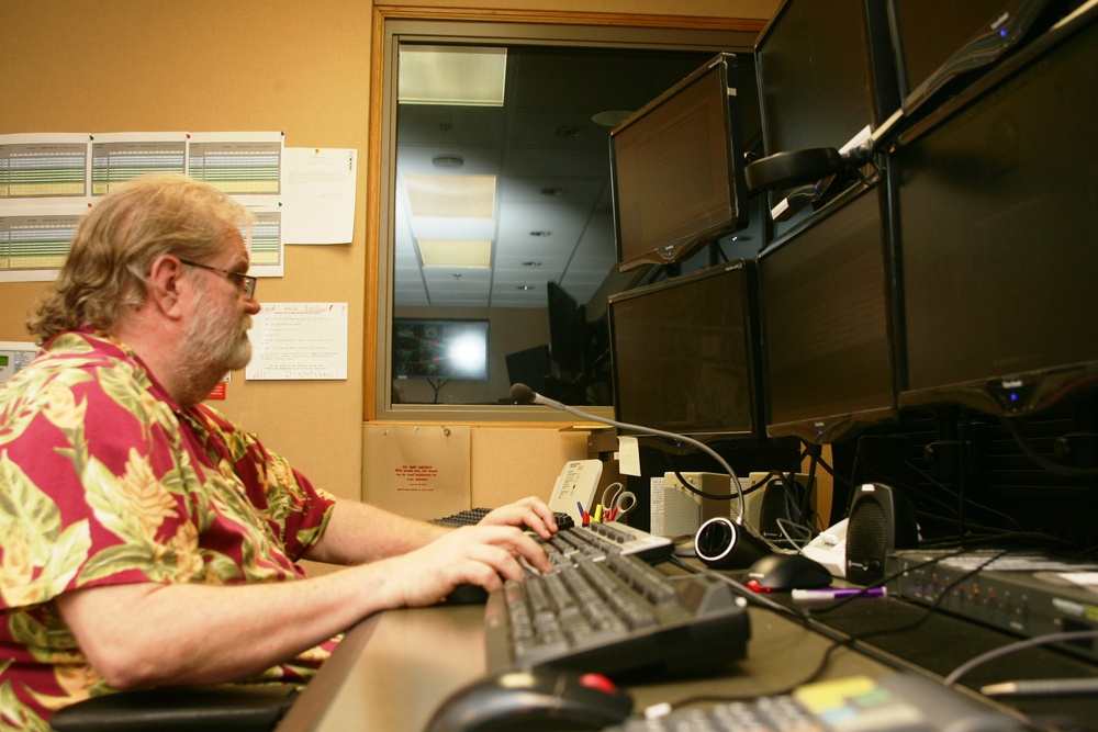 A middle-aged man with glasses and a red floral Hawaiian shirt is seated at a multi-monitor workstation in what appears to be a control room or dispatch center. He is actively typing on a keyboard while facing a bank of at least four dark monitors mounted on adjustable arms. The workspace includes various office supplies, papers pinned to a corkboard wall, and a window looking into an adjacent room.