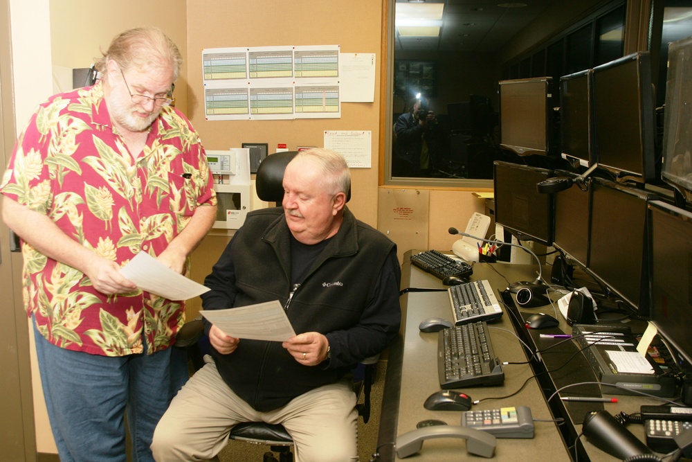 Two older men are reviewing printed documents in what appears to be a dispatch or control room with multiple monitors and keyboards. One man is standing wearing a red Hawaiian shirt, while the other is seated at the workstation wearing a black Columbia fleece jacket. A third person is visible in the background through a window, holding a camera with a flash.
