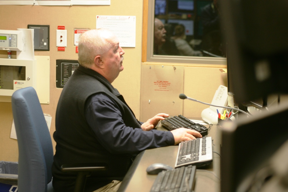 A middle-aged man with gray/white hair is seated at a workstation desk, appearing to be in a control room or security/dispatch center. He is working at a desk with multiple keyboards and a microphone, looking upward or toward a window. Through a window behind him, at least one other person is visible in an adjacent room.