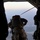 A military paratrooper is captured mid-exit from the open ramp of a military transport aircraft, likely a C-130 Hercules. The soldier is fully equipped with combat gear, a parachute pack, and a rifle, with a static line attached to the aircraft's anchor line cable. The terrain and a distant urban area are visible far below through the open ramp.