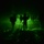 A night vision image showing multiple silhouetted figures in what appears to be a military operation. The green monochromatic imagery is characteristic of night vision optics (NVG/FLIR technology). The figures appear to be moving through an outdoor environment with dust or haze visible in the atmosphere.