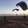 A military parachute operation is captured at dusk on a sandy drop zone, with two parachutists in the final stages of landing under dark ram-air canopies. Ground personnel in military camouflage uniforms are present on the landing zone, which is marked with a bright red panel or streamer used as a wind/landing indicator. The lighting conditions suggest either dawn or dusk operations, consistent with low-visibility tactical training.