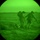 Image captured through night vision optics showing military personnel operating in a dark, open terrain environment. Two standing figures in full combat gear appear to be assisting or moving a third person on the ground. The characteristic circular vignette and green monochrome palette confirm this is a night vision device (NVD) capture.