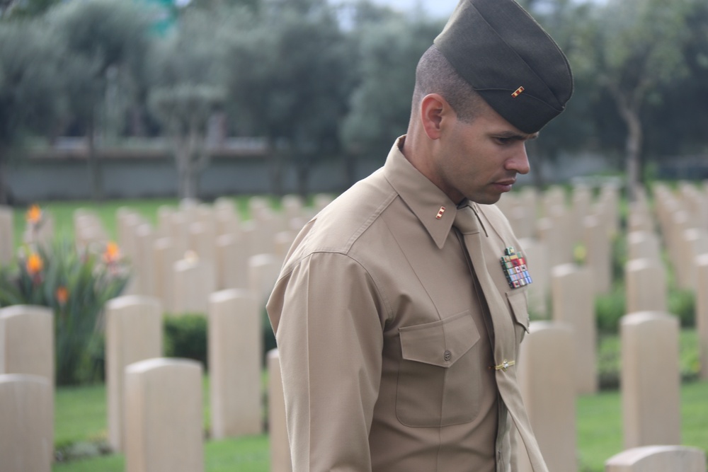 A U.S. Marine Corps officer in service uniform (Charlie uniform) stands with head bowed among rows of white grave markers at a military cemetery. The officer wears a garrison cap with what appears to be a first lieutenant insignia and has several ribbon medals on his chest. The setting suggests a solemn commemorative or memorial visit.