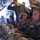 Multiple military personnel in olive/dark green flight suits are seated at workstations inside what appears to be a military aircraft, operating computer equipment and wearing headsets. The crew members display US flag patches on their shoulders, indicating US military affiliation. The scene depicts an active mission environment with personnel monitoring screens and communicating via headsets.