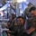 Military personnel in olive drab flight suits are working at a bank of electronic monitoring and control consoles inside what appears to be a military surveillance or command aircraft. Several crew members are wearing headsets and operating workstations with multiple screens and equipment panels. The atmosphere appears relatively relaxed, with some personnel smiling and interacting.