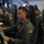 Multiple U.S. military personnel in olive drab flight suits are seated at operational workstations inside what appears to be a military surveillance or patrol aircraft. Each crew member is wearing a headset and working at individual consoles equipped with illuminated green control panels, keyboards, and monitors. American flag patches are visible on their uniforms, indicating U.S. armed forces affiliation.
