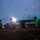 A tiltrotor aircraft, consistent with a V-22 Osprey, is captured at dusk or dawn on an open field with its rotors spinning, visible as green-glowing circular arcs from long exposure photography. Personnel in military gear are visible beneath and around the aircraft, appearing to board or disembark. The scene is illuminated by the aircraft's forward landing light and navigation lights against a dim, cloudy sky.