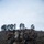 A group of military personnel in full combat gear are gathered on a scrubby hillside terrain, appearing to conduct a field training exercise or tactical operation. Multiple soldiers wearing different camouflage patterns — including MARPAT (Marine pattern) and MultiCam — are clustered together, with at least one appearing to carry or assist another individual on a litter or stretcher. The overcast sky and sparse vegetation suggest a temperate, possibly coastal training environment.