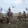 A group of military personnel, appearing to be U.S. Marines based on their distinctive MARPAT camouflage uniforms, are conducting a field training exercise in an open terrain area. The soldiers are in various tactical positions including prone, kneeling, and crouching stances while holding weapons. A V-22 Osprey tiltrotor aircraft is visible in the background sky, suggesting an air assault or insertion exercise.
