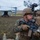 A U.S. Marine in full combat gear is positioned in the foreground holding an M4-style carbine rifle with optical attachments, wearing MARPAT camouflage and a tan MICH helmet with a night vision device mount. Additional Marines are visible in the background, and a V-22 Osprey tiltrotor aircraft is landed on the ground behind them. The scene appears to depict a tactical insertion or post-landing security operation during a training exercise or operational deployment.
