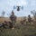 The image shows military personnel in camouflage uniforms taking up defensive positions on open terrain while a tiltrotor aircraft, identifiable as a V-22 Osprey, hovers overhead. The soldiers are armed and positioned in prone and kneeling stances, suggesting a tactical insertion or security exercise. The setting appears to be a training environment based on the controlled, photogenic composition.