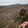 A military service member in full combat gear and camouflage uniform is conducting a field training exercise among sparse brush and scrubland. A training mannequin is visible on the ground nearby, indicating a casualty care or combat scenario drill. The setting appears to be a military training range with rolling hills in the background.