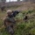 The image shows military personnel conducting what appears to be a field training exercise in an open, grassy terrain. A foreground soldier in MARPAT (Marine pattern) camouflage is in a kneeling firing position aiming a rifle equipped with optics and accessories. Two additional soldiers are visible in the background in tactical stances.