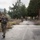 A group of military personnel in camouflage uniforms are conducting what appears to be a patrol or training exercise along a paved road near an abandoned or deteriorating structure. The lead soldier in the foreground is carrying an assault rifle with an underslung grenade launcher attachment and wearing full combat gear including a helmet with a mounted device. The setting appears to be an overgrown, semi-abandoned military or industrial facility surrounded by trees and hills.