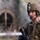 A U.S. Marine in full combat gear is photographed in a close-up portrait shot. He is wearing MARPAT woodland camouflage uniform, a tan MICH/ACH combat helmet with a night vision goggle mount, and a tactical headset with boom microphone. The background is blurred but shows what appears to be a stone archway or tunnel structure in an outdoor environment.