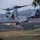 A U.S. Marines MV-22 Osprey tiltrotor aircraft is captured in low-altitude flight or hover, with rotors in vertical/helicopter mode. The aircraft bears markings identifying it as belonging to VMM-266 squadron with tail number 8636 and modex '00'. The background shows a hilly, semi-rural landscape with scattered buildings and vegetation.