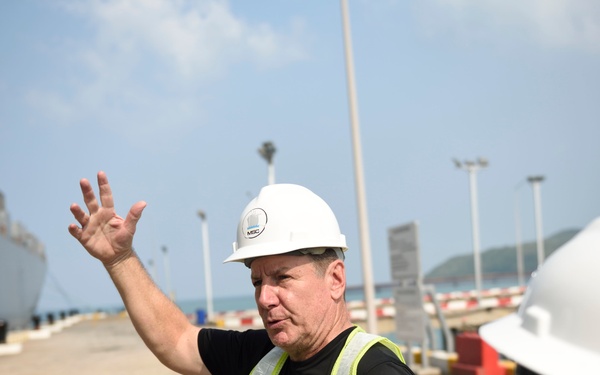A man wearing a white hard hat labeled 'MBC' and a high-visibility yellow safety vest is gesturing with his raised hand, apparently briefing or directing two other individuals at a port or industrial dock facility. A large naval or cargo vessel is visible in the background to the left. The setting appears to be an active working waterfront area.