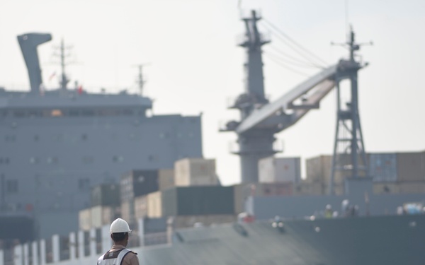 A port worker wearing a white hard hat and reflective safety vest stands on a dock, observing a large gray vessel — likely a military or naval auxiliary ship — approaching or docking. The ship is loaded with shipping containers on its deck and features a prominent superstructure with cranes and communication masts. A second large vessel is partially visible in the background to the left.